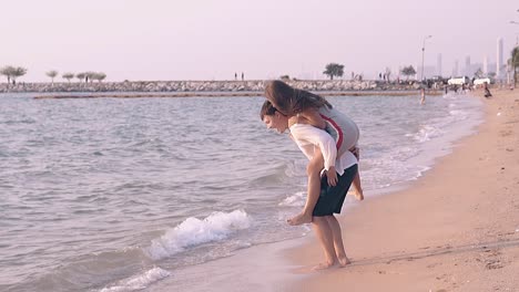 una chica alegre salta sobre un chico de vuelta en la orilla del mar en cámara lenta