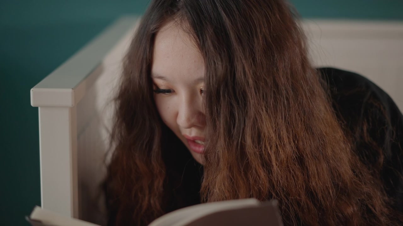 Close-up of young girl reading book with steady downward gaze and slightly open mouth suggesting soft speech; calm expression, quiet focus, and curious attention create intimate home mood