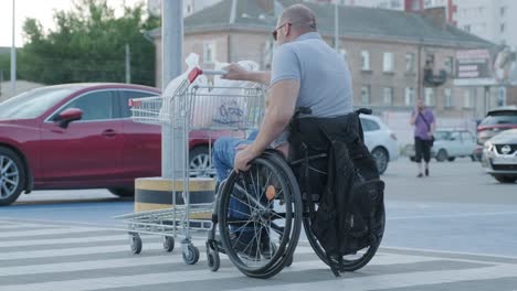 man with disabilities in wheelchair pushing cart in front of himself at supermarket parking