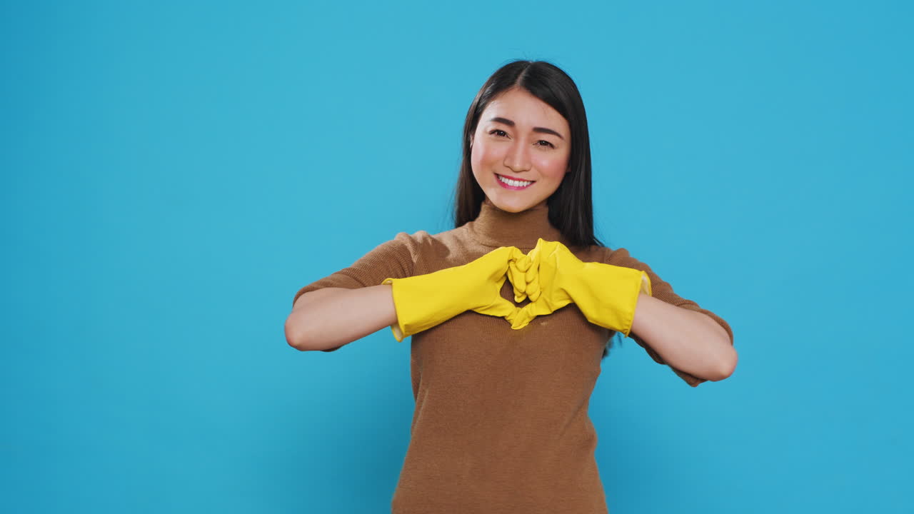 Happy asian maid wearing yellow gloves while doing heart shape in front of camera after finishing cleaning houses