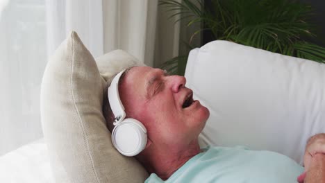 caucasian senior man wearing headphones singing while lying on the couch at home