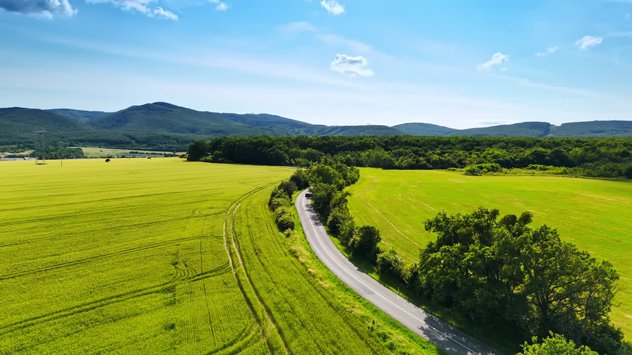 Driving through winding fields. A black car travels down a winding road surrounded by lush green fields and distant mountains on a sunny day