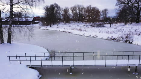 small city park with bridge over frozen lake water in winter season during snowfall, aerial view