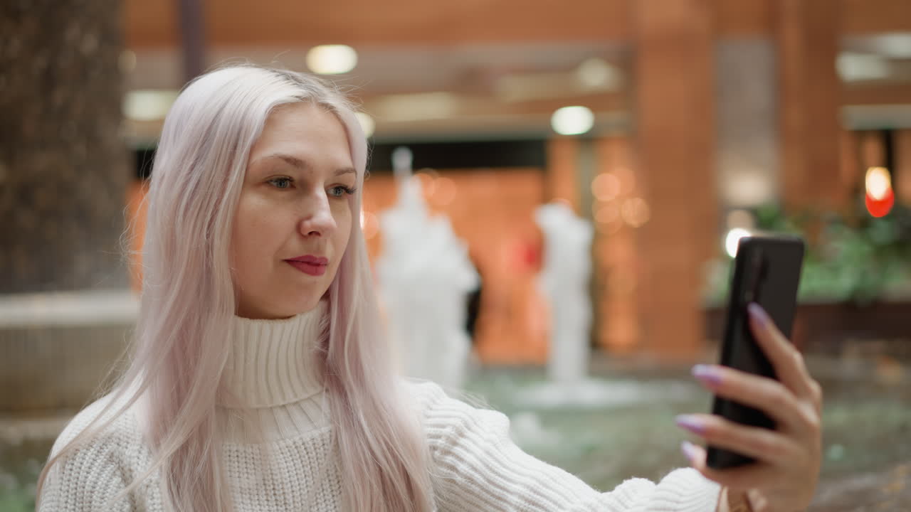 Urban dweller sits on fountain bench in mall, adjusts hair and poses with mobile phone for selfie, smiles into camera as water jets sparkle in blurred retail interior under warm lights