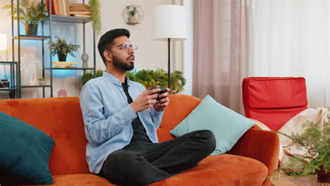 joven indio usando un controlador de joystick jugando videojuegos ganando juegos en línea en el sofá de casa