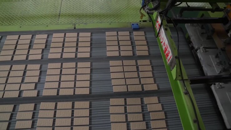 Pressa di trasferimento automatica che si solleva sopra un ampio letto di piastrelle pressate color marrone chiaro su una macchina di produzione verde