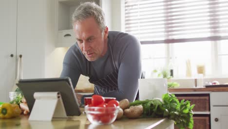 Hombre-Caucásico-Feliz-Usando-Delantal,-Parado-En-La-Cocina,-Cocinando-La-Cena-Y-Usando-Una-Tableta