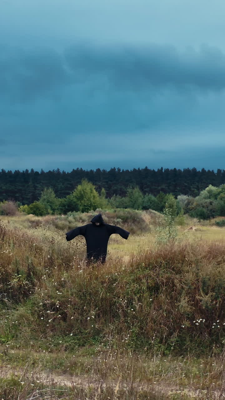 Dark scary figure on the field. Black death in mantle standing with outstretched arms among summer field under evening sky. Vertical video
