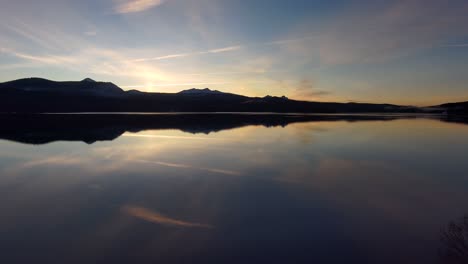 Drone-through-trees-over-reflective-lake-at-sunrise-with-mountains,-silhouette,-60fps