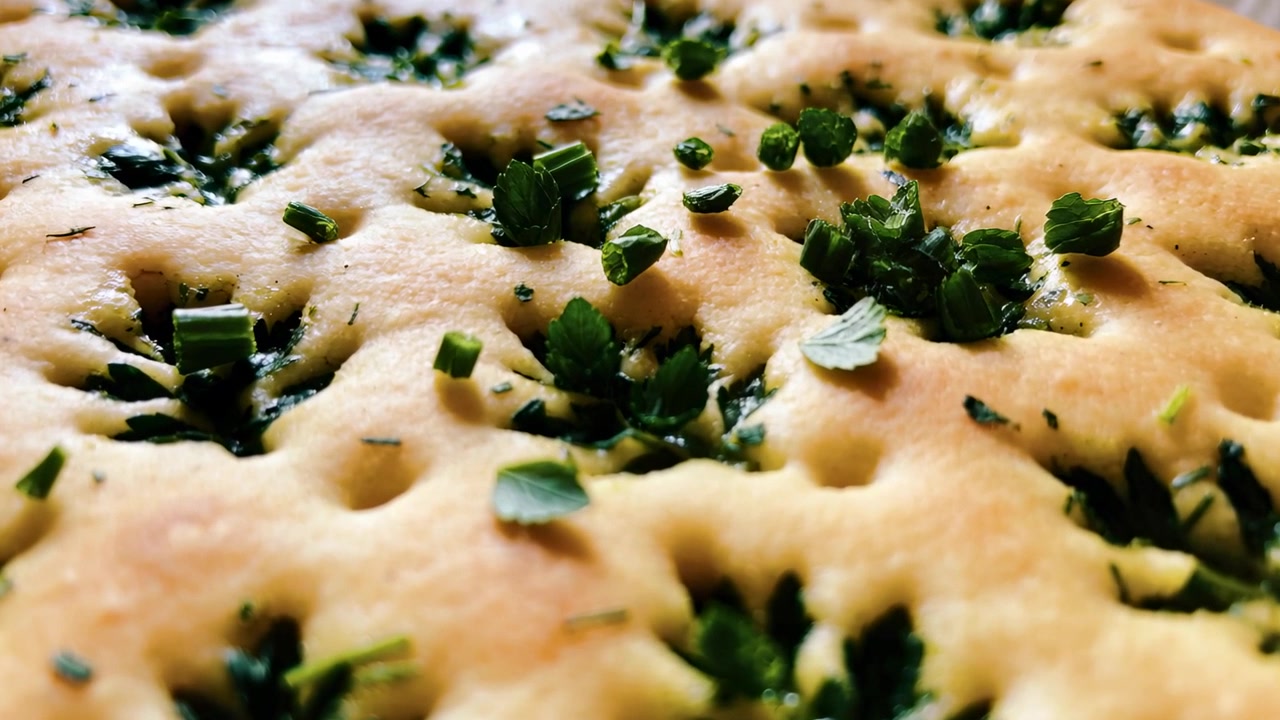 Telecamera che sposta la messa a fuoco riprendendo una focaccia alle erbe con le erbe che si depositano nelle fossette su un vassoio in cucina.