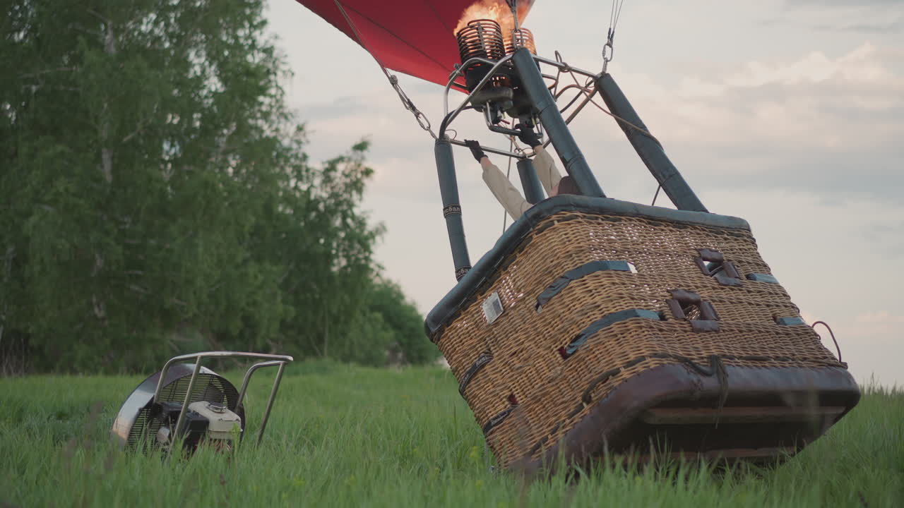 pilot operating burner to lift hot air balloon basket and inflate envelope over grass field at dusk, gripping control frame as vibrant canopy rises under calm twilight sky in preflight