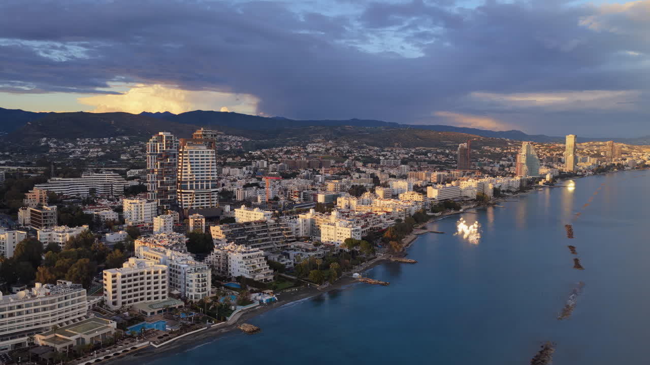 Vista aérea de drone de arranha-céus modernos em Limassol iluminados pela luz quente da hora dourada com montanhas ao fundo.