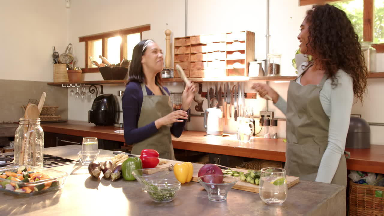 Multiracial female friends dancing joyfully in kitchen while preparing meal together, at home