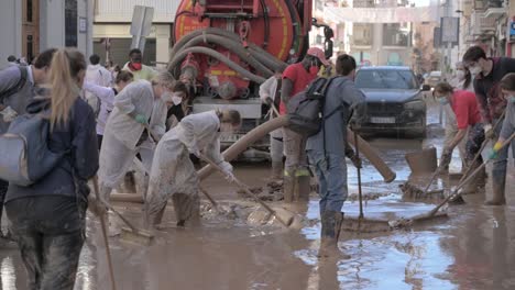 a group of volunteers working together to clean muddy streets following a heavy flood in valencia. people use shovels and brooms to remove debris while a truck sucks up the mud