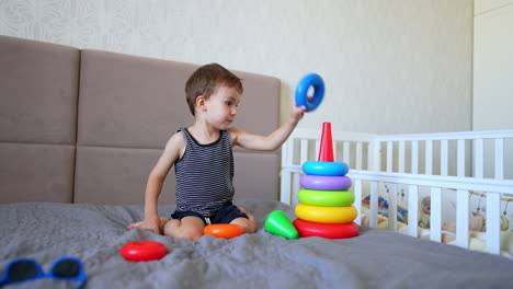 lovely small caucasian kid sitting on double bed playing with toy. cute kid building a colorful pyramid.