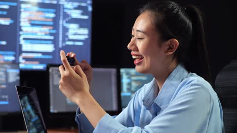 vista lateral de cerca de una programadora asiática mirando un teléfono inteligente celebrando el éxito mientras escribe código en una computadora portátil usando múltiples monitores que muestran la base de datos en los escritorios de la oficina