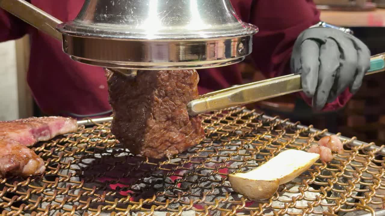 Gloved chef grills wagyu steak on open charcoal grill using metal tongs and smoke dome