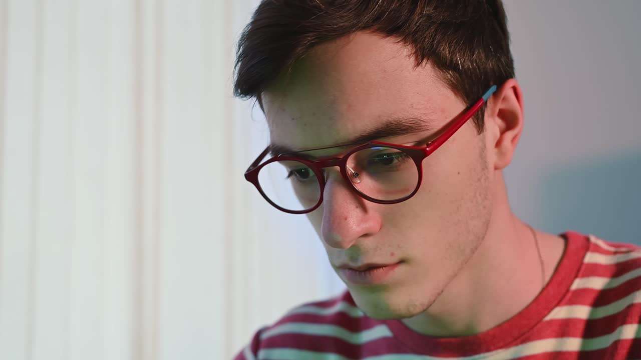 Young pimply man in red glasses and pink-striped sweater writing codes on a green page in his computer. Concentrated programmer. Reflection in the glasses. Side view. Close up