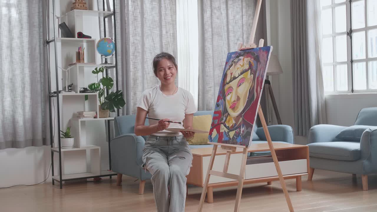 Asian Artist Girl Sitting On Round Wooden Chair Without Backrest Holding Paintbrush Mixed Colour And Smiling After Finish Painting A Girl On The Canvas
