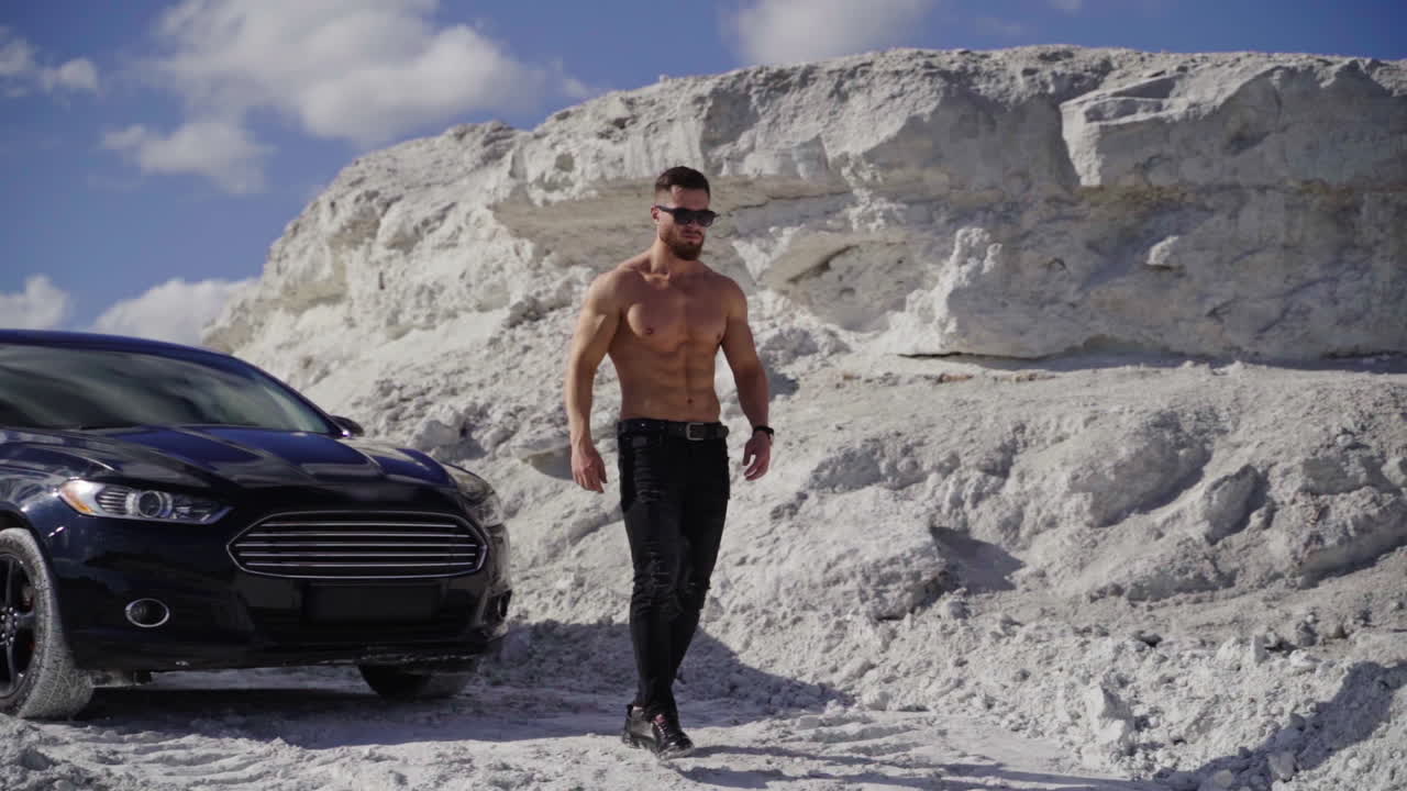 Handsome man near the black car. Shirtless guy in black sunglasses with sportive body walking in white mountains in a summer day. Slow motion.