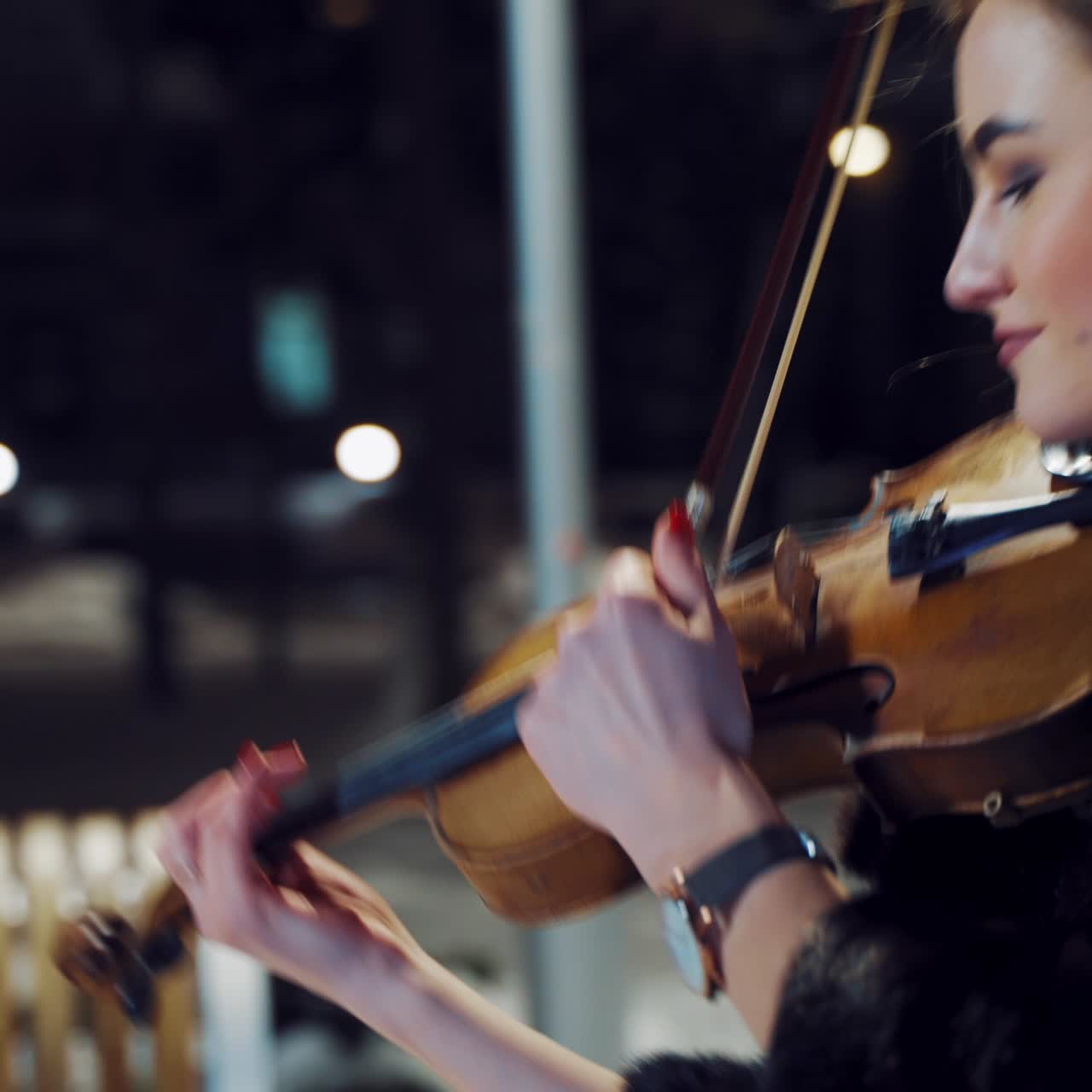 A pretty girl stands in the middle of the square and holds the violin with one hand, the bow with the other hand and performs the composition in the evening. Close-up.