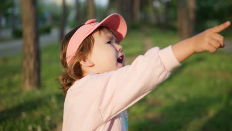 Aufgeregtes-3-jähriges-Kleines-Mädchen-Zeigt-Mit-Dem-Finger-Auf-Etwas-Interessantes-Und-Redet,-Während-Es-Bei-Sonnenuntergang-Im-Grünen-Baumpark-Steht