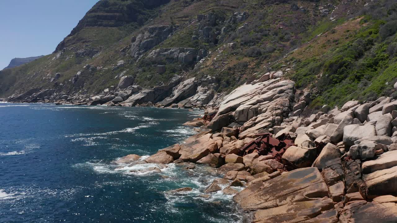 Aerial view moving along steep rocky shoreline littered with shipwreck debris in Cape Town