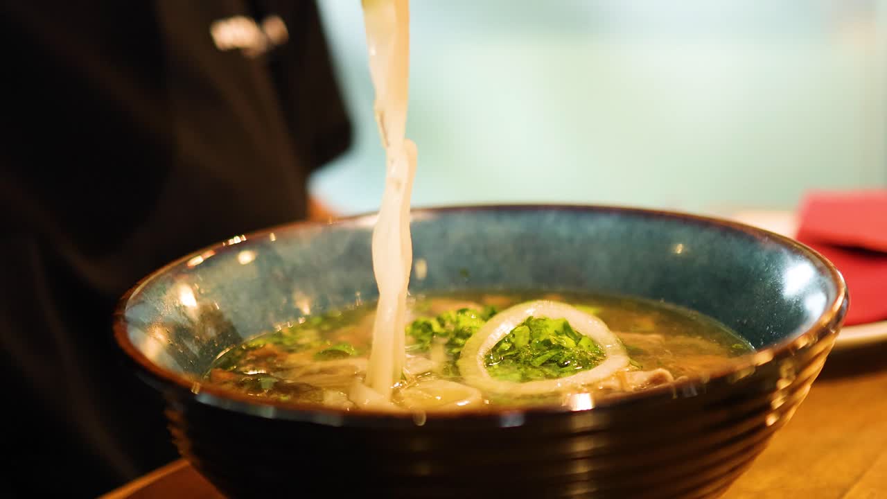 Chopsticks lift noodles from a steaming bowl of pho in a cozy Bangkok eatery, highlighting the vibrant culinary scene