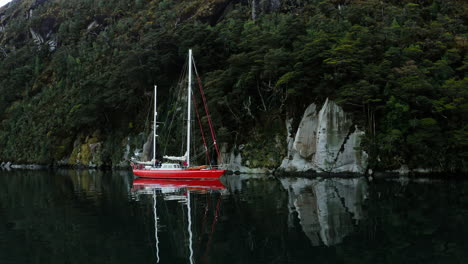 red sailboat drifts still in glassy fjord waters with symmetrical forest reflections, medium orbit aerial