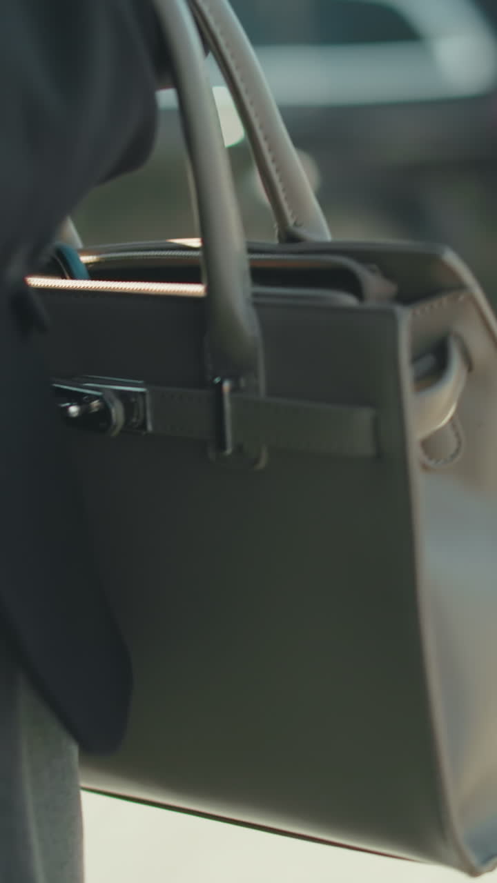 Close up partial view of lady in business outfit carrying handbag walking by on city sidewalk, with blur view of moving car in background