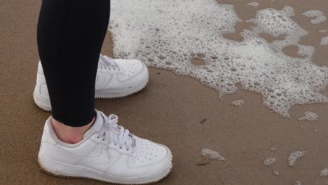 Girl-in-activewear-and-white-trainers-stands-on-the-sandy-beach-as-foam-from-crashing-ocean-waves-flow-up-to-her-toes-and-fall-back-in-slow-motion-on-dull-grey-day
