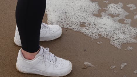 girl in activewear and white trainers stands on the sandy beach as foam from crashing ocean waves flow up to her toes and fall back in slow motion on dull grey day