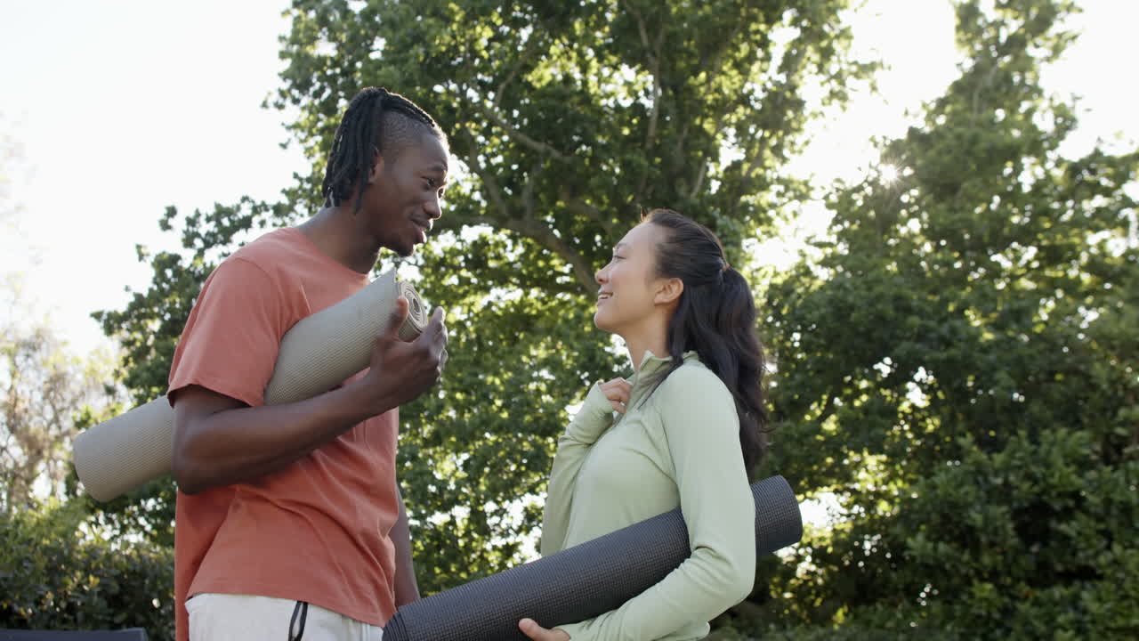 Happy diverse couple holding yoga mats talking in sunny garden, slow motion