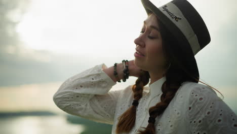 un primer plano de una mujer con un sombrero negro y un vestido blanco, sonriendo satisfecha bajo un cielo nublado. su mano descansa cerca de su cuello, adornada con pulseras de cuentas