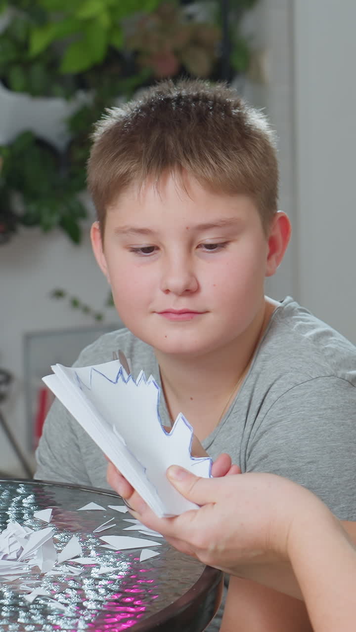 백인 남자아이가 테이블에서 종이 오리기를 보고 있어요. 어른의 손이 가위를 잡고 눈송이 패턴을 만들도록 안내하고, 실내 식물이 배경에 있으며, 차분한 튜토리얼 분위기 속에서 아이는 집중하여 관찰하고 부드러운 지시가 이어집니다.