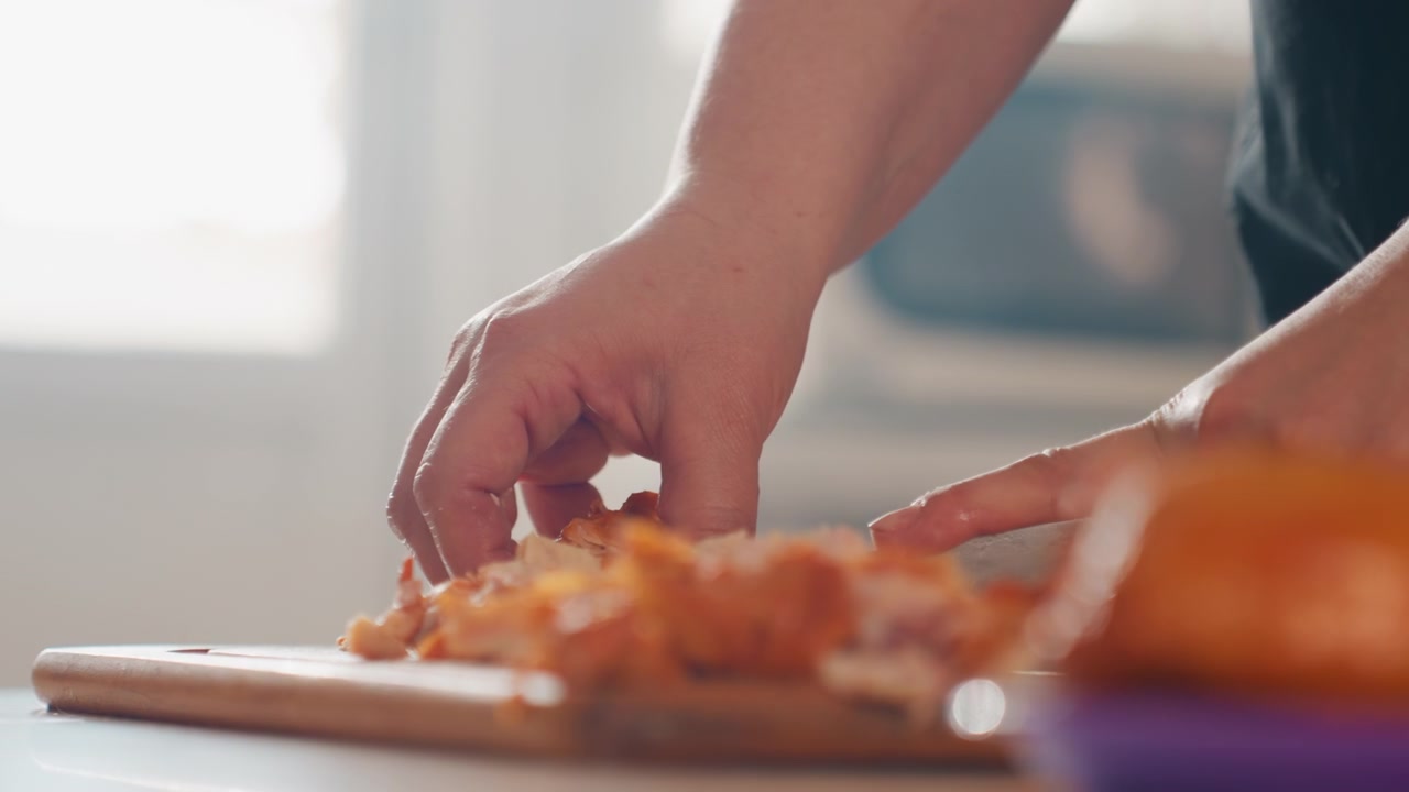 Close-up of hands pulling apart cooked meat on wooden board beside bun in sunlit kitchen, with knife steadying food and visible finger movement during homemade meal preparation, with hand, shredding