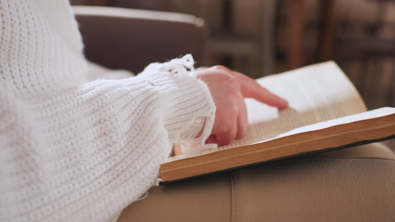 Close view of female hands flipping through textbook while seated and pointing at page with index finger, dressed in cozy knit sweater, highlighting focused study moment in calm indoor setting
