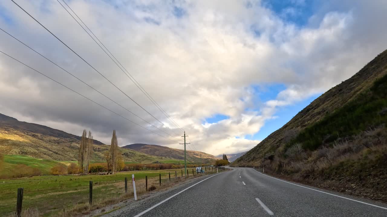 A serene drive through Wanaka's picturesque landscape, featuring winding roads, lush hills, and dynamic cloud formations
