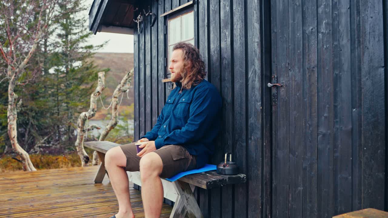 A Caucasian Man Seating Outside The Wooden Cabin Drinking Coffee. Static Shot