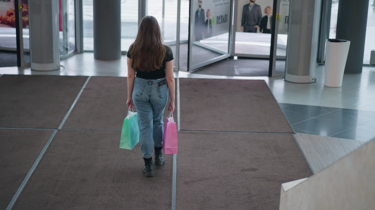 mujer llevando bolsas de compras caminando hacia la salida en un centro comercial moderno, persona que pasa, puertas giratorias en movimiento, traje elegante, botas casuales, experiencia de compras urbanas con iluminación brillante