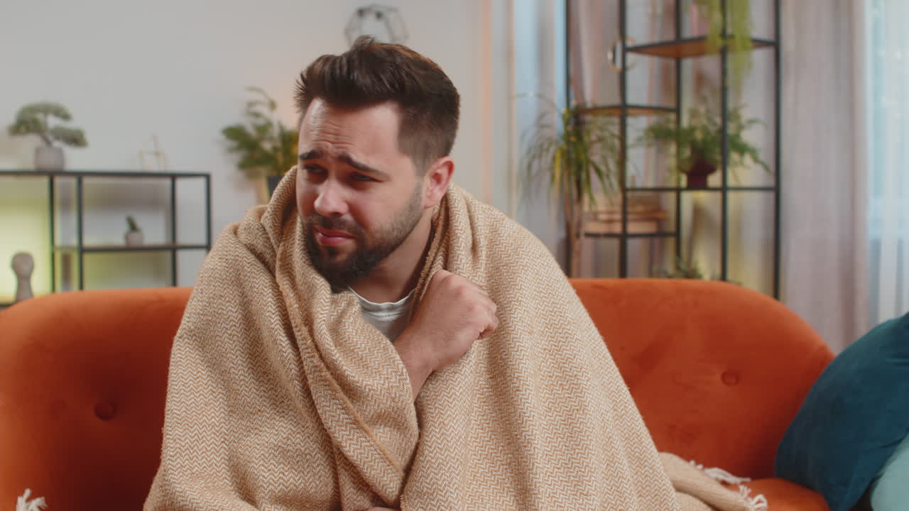 joven caucásico enfermo cubierto de una manta toser mientras está sentado en el sofá en la sala de estar