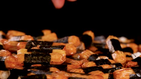 A hand reaches into a pile of roasted rice crackers wrapped in seaweed, illuminated by dramatic studio lighting against a black background, captured in smooth motion