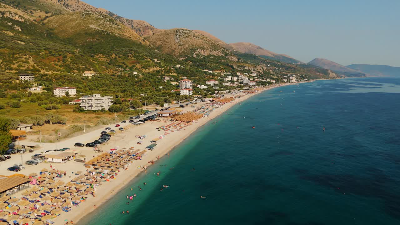 A crowded beach with turquoise water and mountain landscape in the distance, aerial view