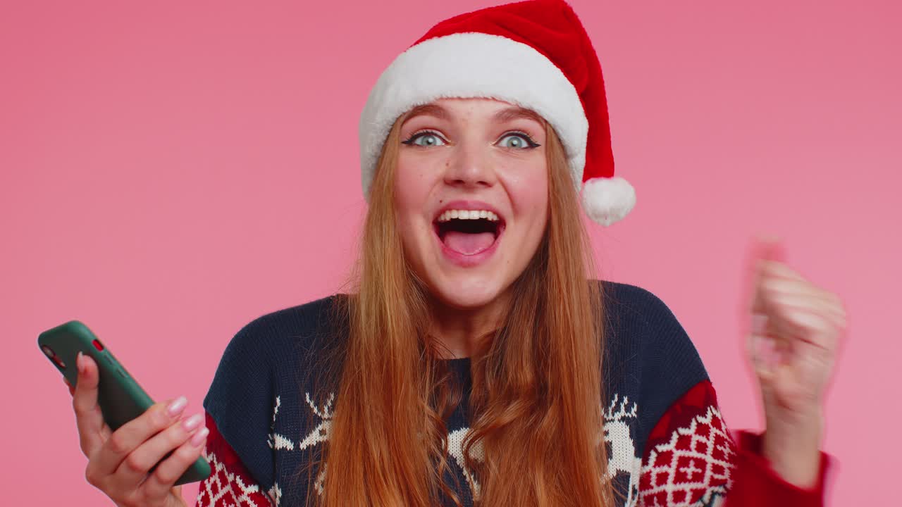 스마트폰 디스플레이를 보고 있는 세련된 크리스마스 스웨터를 입은 여성은 진심으로 기뻐하며 성공을 기원합니다