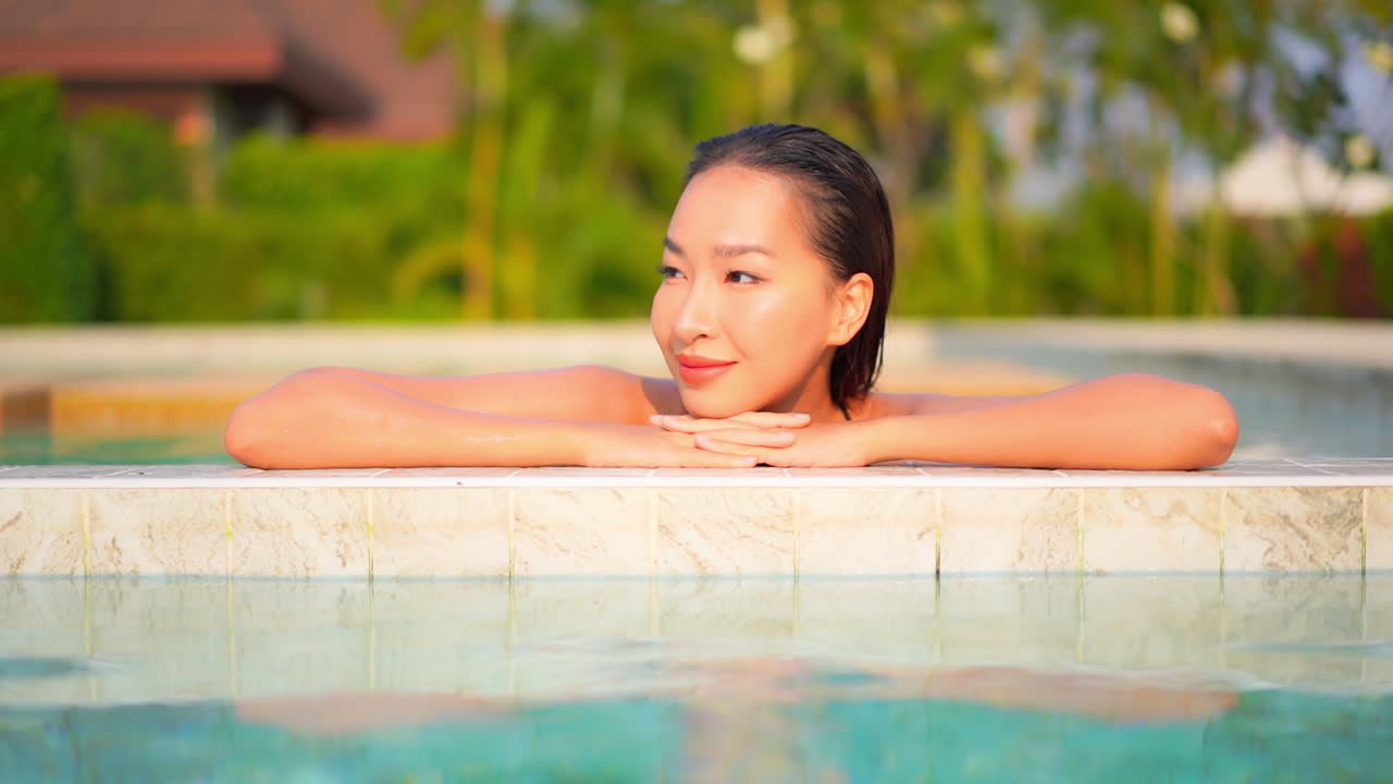 수영장 가장자리에 기대어 있는 매력적인 젊은 여성이 팔에 머리를 얹고 눈을 감습니다.