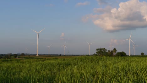 Windkraftanlagen-Schießen-Aus-Einem-Ackerland,-Während-Die-Sonne-Untergeht,-Während-Lastwagen-Vorbeifahren-Und-Staubwolken-Erzeugen,-Saubere-Alternative-Energie-In-Thailand-Und-Auf-Dem-Südostasiatischen-Festland