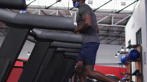 fit african american man with face mask around his neck running on treadmill in the gym