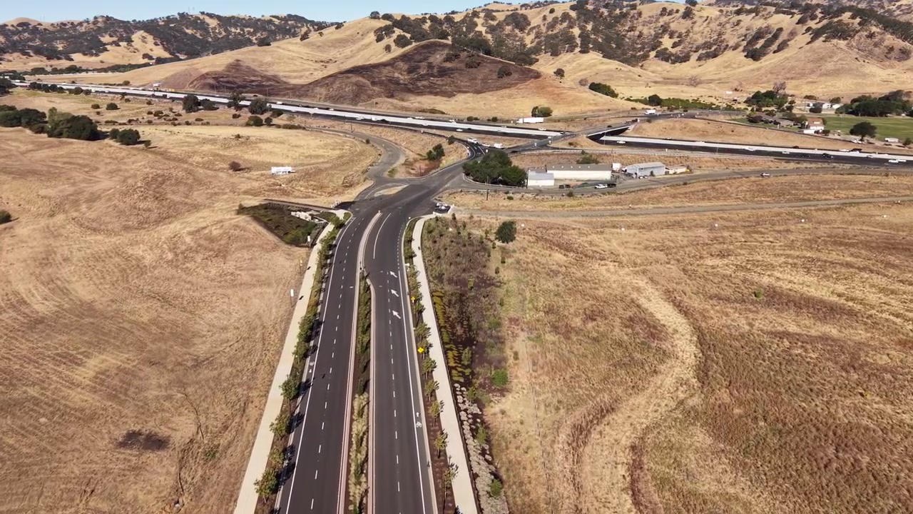 Riprese aeree con drone che volano in avanti lungo una strada nel Lagoon Valley Park, Vacaville, California, mostrando il paesaggio arido e un incrocio autostradale distante con colline ondulate marroni.