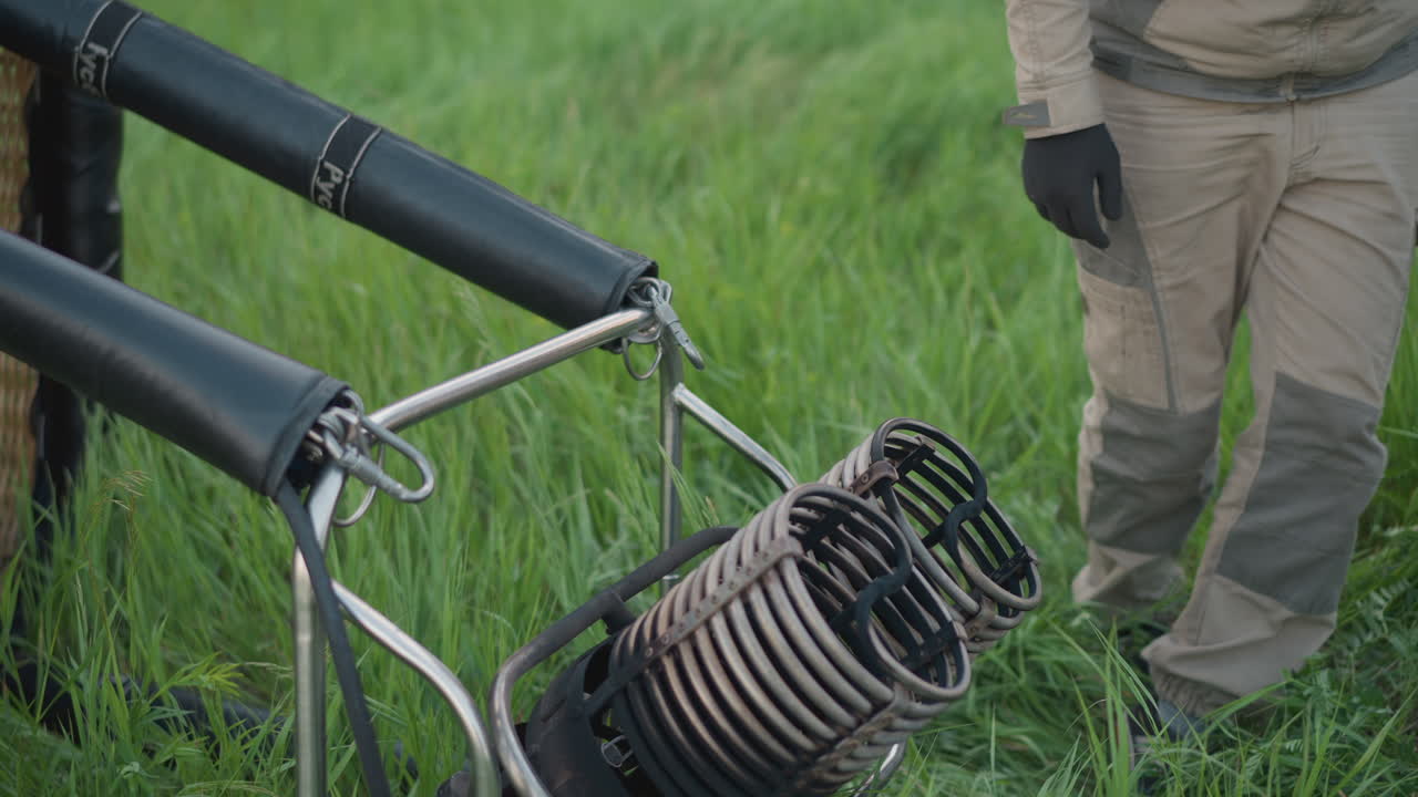 metal burner rests on grassy field during preflight as colleague secures burner assembly then stands, walks away beside uninflated balloon envelope under cloudy sky in rural setting