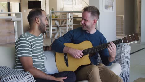 pareja de hombres gay multiétnicos sentados en el sofá uno tocando la guitarra